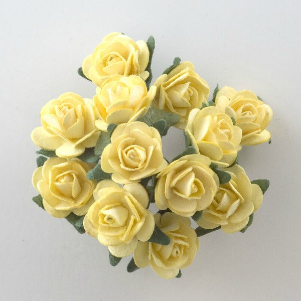 A cluster of Lemon Paper Tea Roses Bunch of 12 from Perfect Tables, featuring soft, delicate petals and green leaves with visible stems, are arranged in a circular formation on a light, neutral background. These small, pale yellow roses have a slightly curled texture, making them perfect for craft projects.