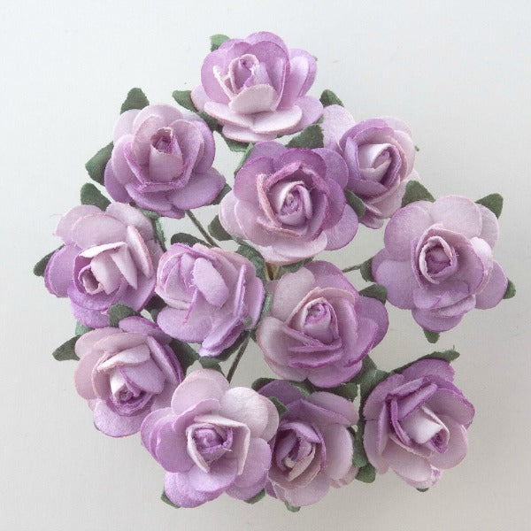 A bouquet of light purple Lilac Paper Tea Roses Bunch of 12 from Perfect Tables, artfully arranged in a circle and captured from above. The roses, resembling delicate paper tea roses with their layered petals, are set against a plain white background. The green leaves partially visible among the flowers enhance the stunning colors of this craft project masterpiece.