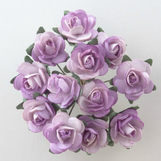 A bouquet of light purple Lilac Paper Tea Roses Bunch of 12 from Perfect Tables, artfully arranged in a circle and captured from above. The roses, resembling delicate paper tea roses with their layered petals, are set against a plain white background. The green leaves partially visible among the flowers enhance the stunning colors of this craft project masterpiece.